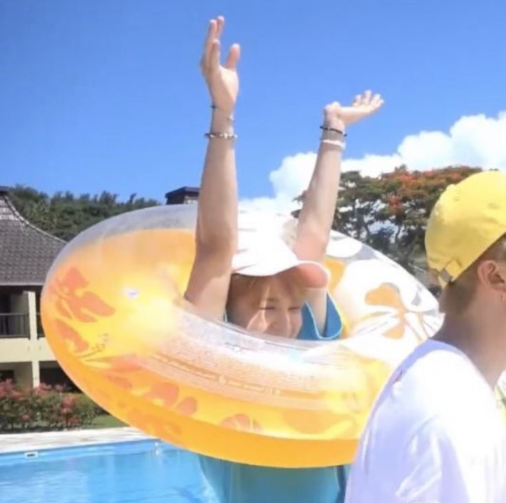 a tiny, summery blonde jimin emerging as if newly born from an inflatable yellow pool ring. his arms are held straight up in the air, most of his smiling face free from the ring, and he has smiling crescent eyes, a baby pink cap, and a blue t-shirt on. 