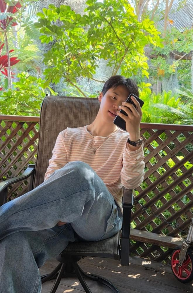 candid jimin photo by hobi. jimin in jeans and a long sleeve top in an outdoor chair with greenery behind him. 