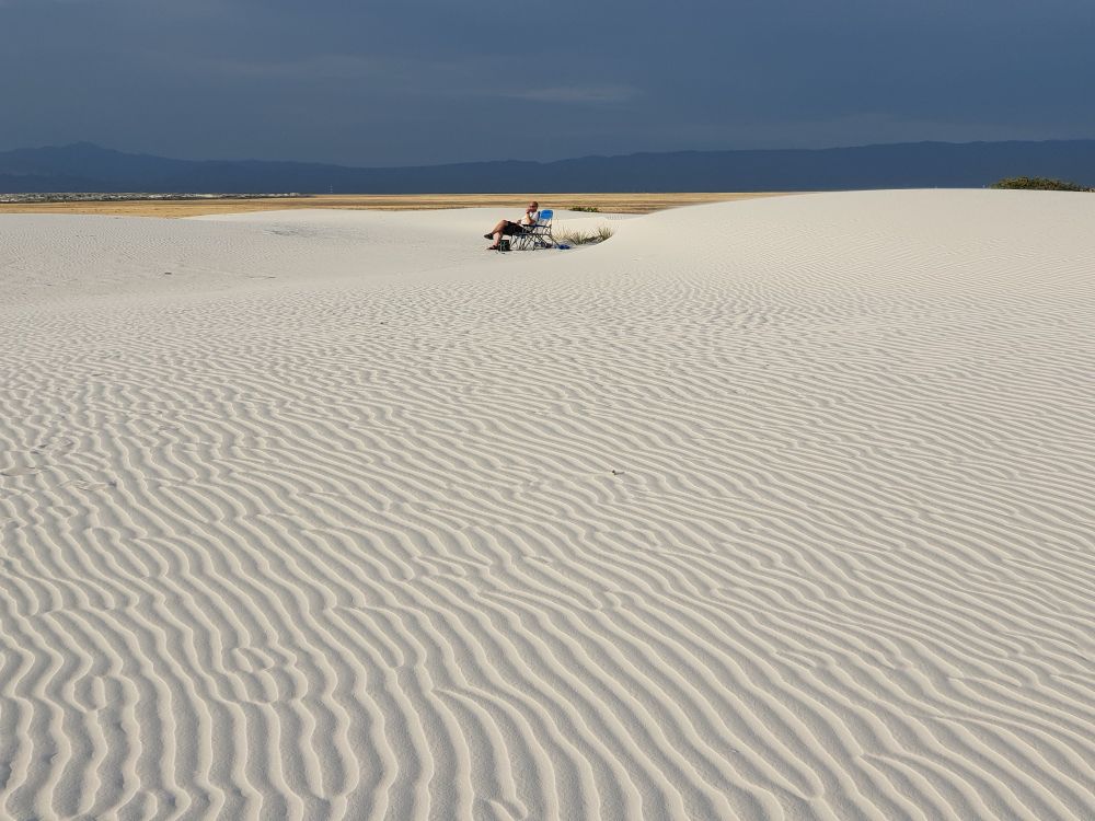 Eine weiße Kalksandfläche, die von Rillen durchzogen ist, die der Wind erschaffen hat. Weit hinten sitzt ein einzelner Mensch, darüber der wolkenlos blaue Himmel.
