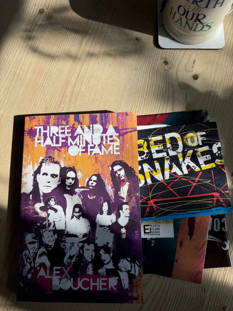 A copy of the book 'Three and a Half Minutes of Fame' by Alex Boucher on a wooden table, showing the two CDs, poster, and stickers it arrived with. The sun is casting some nice shadows across the picture. A cup of tea sits nearby, ready to kick start the day.