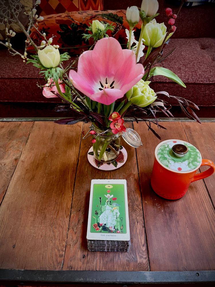 A jar of tulips and anemones on a wooden coffee table with a red tea mug and a tarot card: The Empress