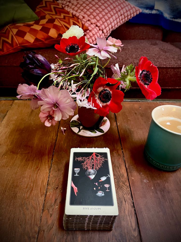 Wooden table on which is a small vase of flowers - red anemones, pink jasmine and ranunculus, a deep purple “Vincent Van Gogh” tulip- and a tarot card: the Five of Cups