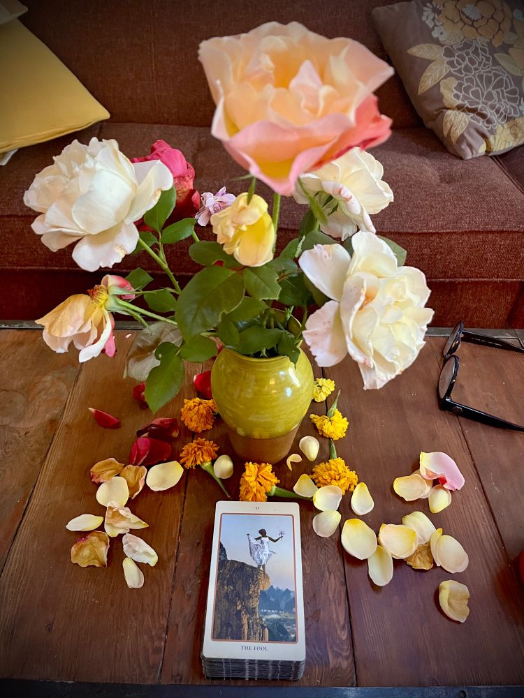 A wooden table bearing a vase of wilted roses, fallen petals, and a tarot card: The Fool