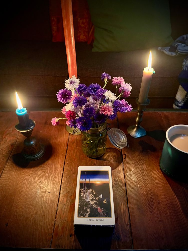 Candles on a table in a dark room with a jar of cornflowers and a tarot card: the Three of Wands