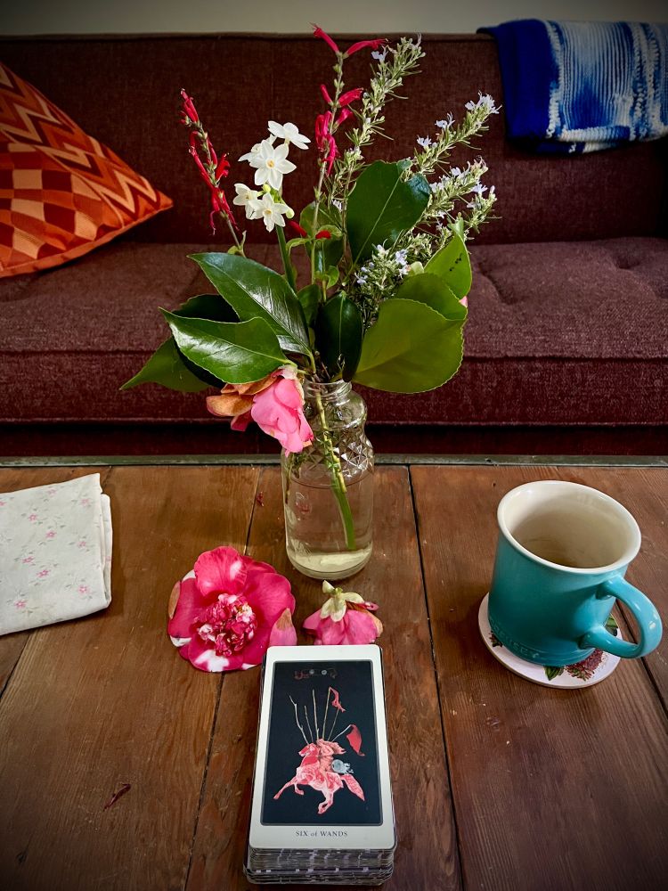 wooden table, bottle of flowers with fallen camellia blooms all around it, a teacup, and a tarot card: the Six of Wands