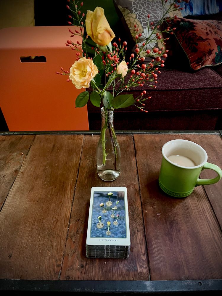 On a wooden coffee table, a green mug of tea and a vase of small orange roses, and a tarot card: Seven of Coins 