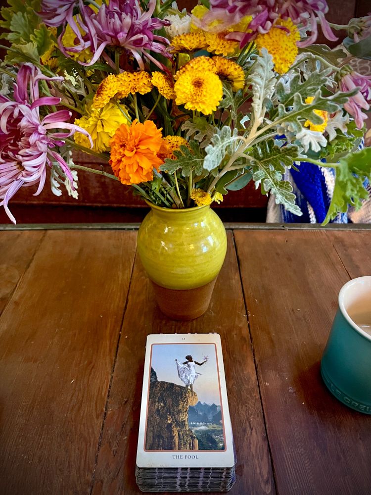Wooden coffee table, ceramic vase glazed chartreuse green; inside, a bouquet of winter flowers —purple mums, yellow marigolds, silver Dusty Miller— and in front of them a tarot card: The Fool