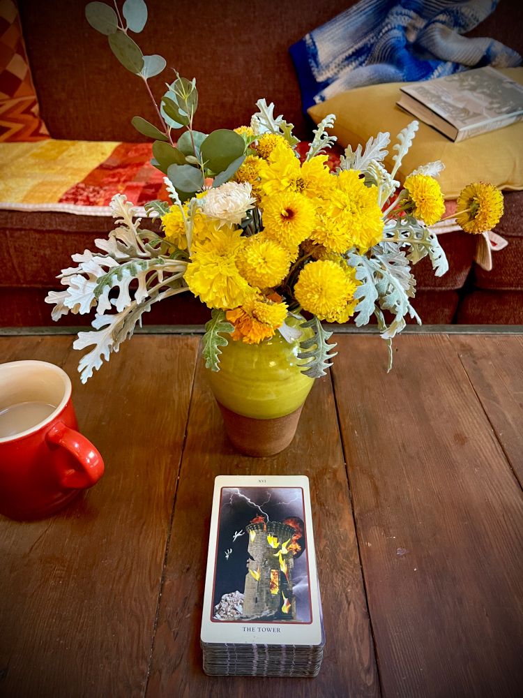 Wood coffee table, yellow ceramic vase, bouquet of late-season yellow flowers and grey foliage, red coffee mug, and a tarot card: The Tower 