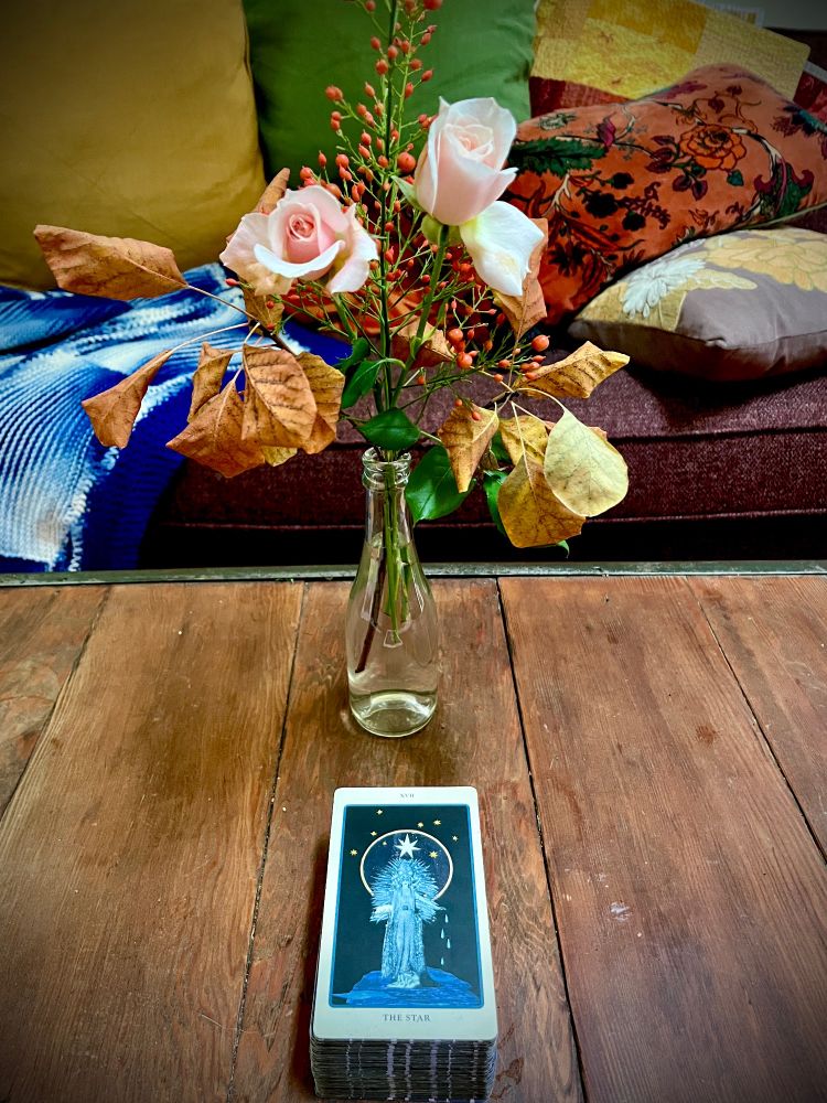 Wooden table with couch and multicolored pillows in the background, foregrounding a jar of pink roses and orange smoke-bush leaves, and a tarot card: The Star