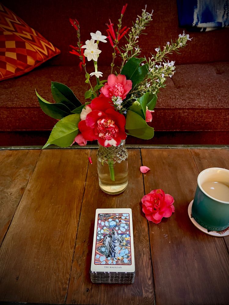 a jar of camellia, rosemary, and salvia flowers on a wooden table: a coffee mug; and a tarot card: The Magician