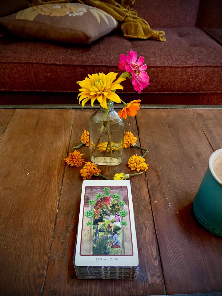 a wood table bearing a small jar of orange and pink flowers, surrounded by a circle of drying marigolds, and a tarot card: the Ten of Coins