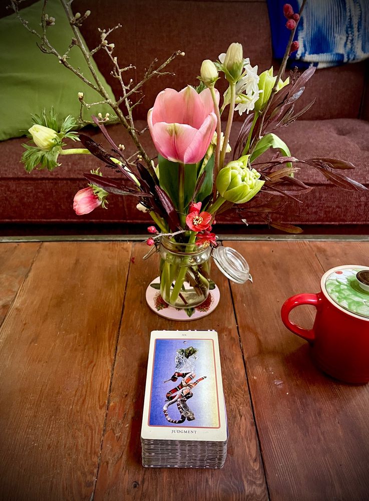 A jar of tulips and tree blossoms on a wooden table alongside a red mug and a Tarot card: Judgment