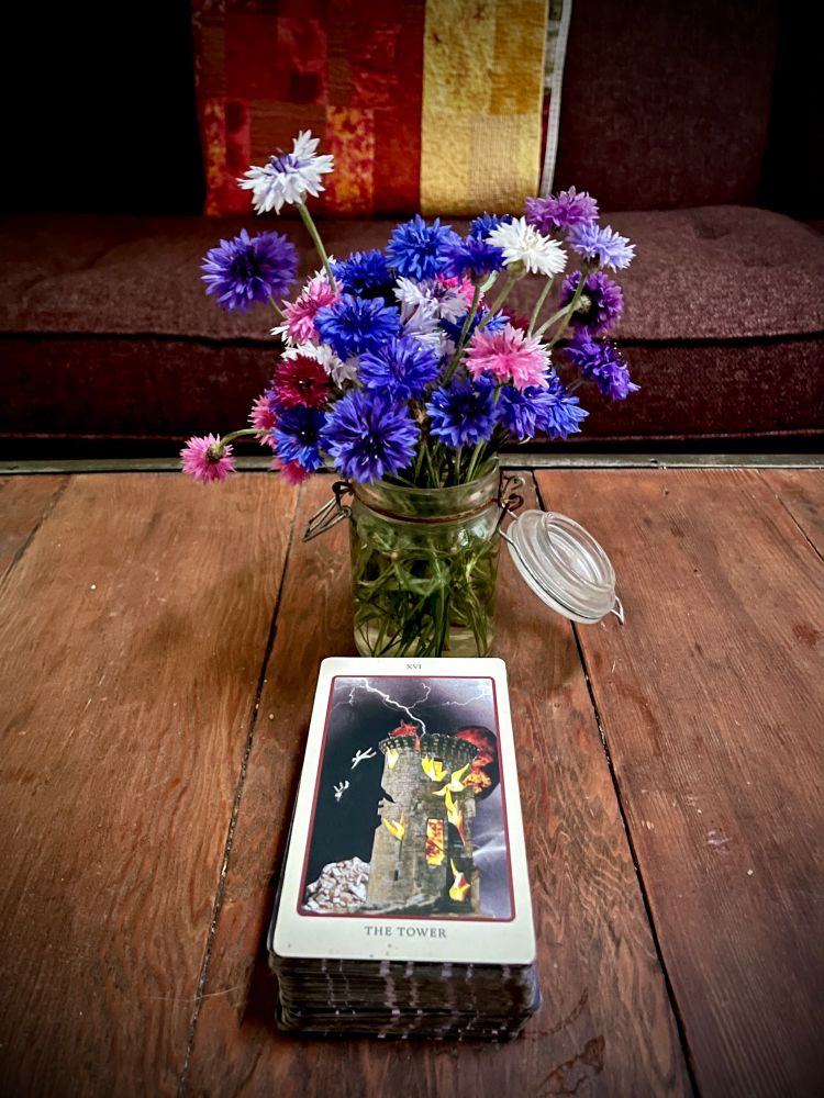 Wooden table in front of a red couch; on the table is a small jar full of blue bachelor’s button flowers and a tarot card: The Tower