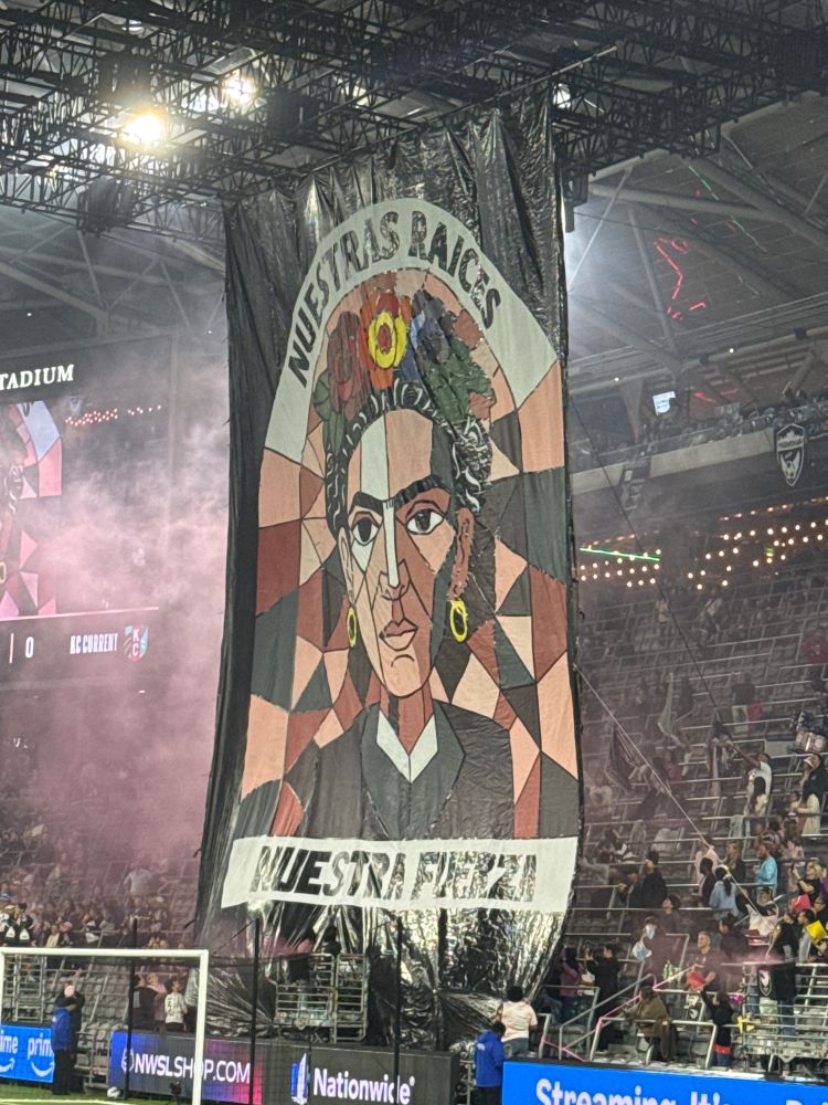 Huge banner featuring a mosaic-style portrait of Frida Kahlo in shades of pink, mauve, and black. It reads “Nuestra raices, Nuestra Fuerza.”