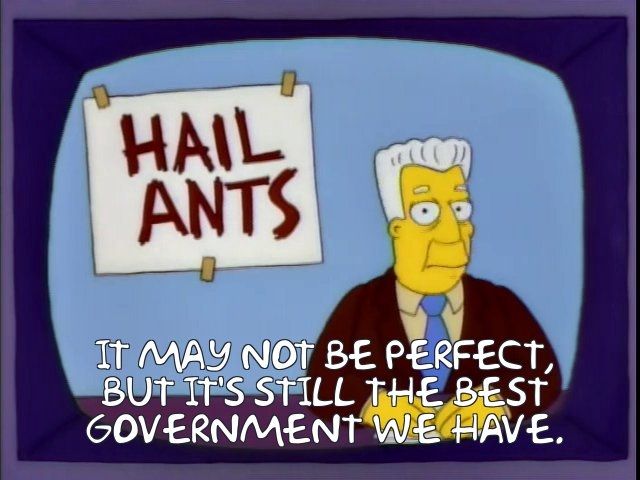 A Simpsons screenshot of Kent Brockman at the news desk with a taped up sign behind him that says "HAIL ANTS" and the caption "IT MAY NOT BE PERFECT, BUT IT'S STILL THE BEST GOVERNMENT WE HAVE." 