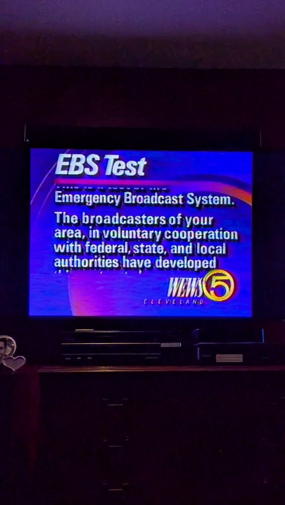 A TV displaying the old Emergency Broadcast System Test with the text, "Emergency Broadcast System. The broadcasters of your area, in voluntary cooperation with federal, state, and local authorities have developed" and the WEWS 5 Cleveland logo from that era. 