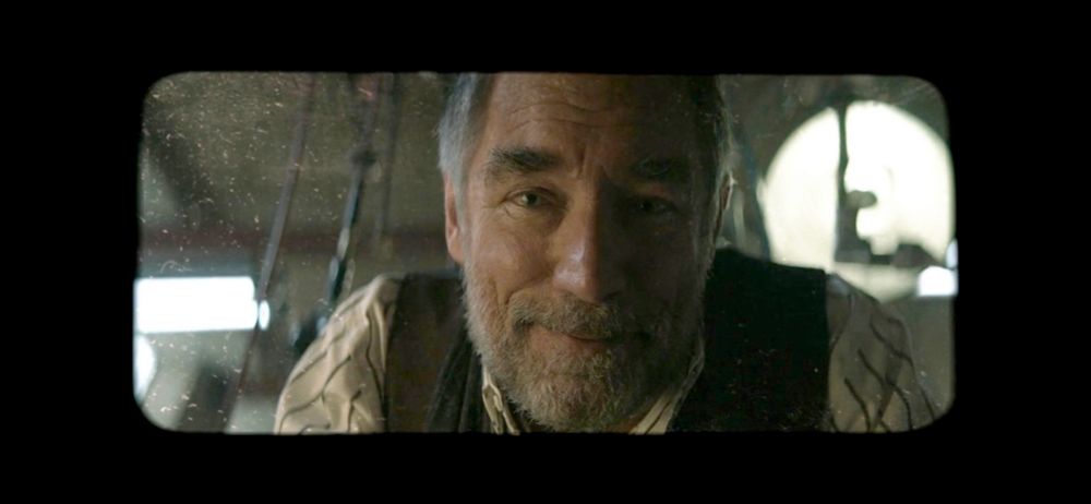 A screenshot of Niles Caulder from the Doom Patrol TV show adaptation. The shot has a thick black border, with a rectangular lens of sight, implicating limited vision. Niles is looking at the viewer, with a bittersweet smile. There is dust on the lens, and behind Niles is machinery and work lights.