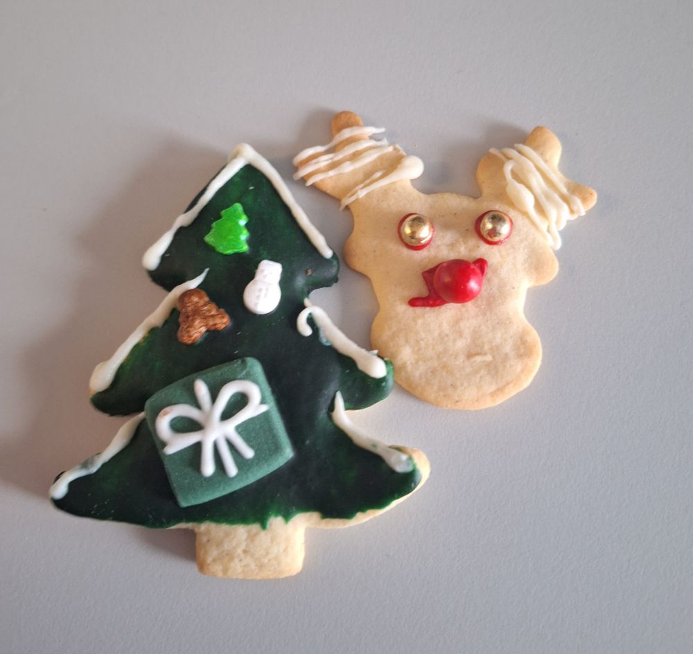 Zwei weihnachtlich verzierte Kekse auf einem hellen Untergrund. Links ein grüner Weihnachtsbaumkeks mit weißem Zuckerguss, kleinen Dekorationen und einem Geschenkmotiv. Rechts ein Rentierkeks mit roter Nase, goldenen Augen und weißem Zuckerguss an den Ohren.