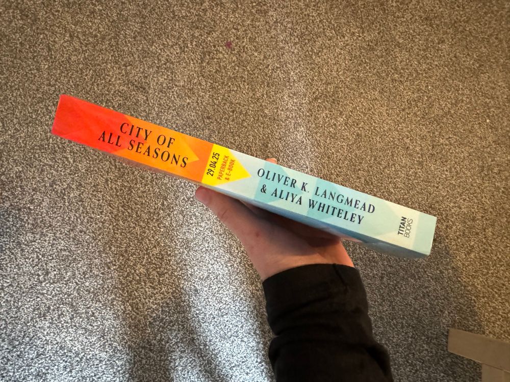 A hand holding a copy of an ARC of A City Of All Seasons by Aliya Whiteley and Oliver K Langmead. The book is held so the spine is uppermost and facing the camera. 