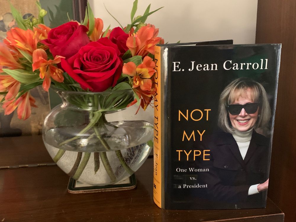 short bouquet of red and orange flowers sits next to E. Jean Carroll's memoir NOT MY TYPE, photo of her in a dark jacket over a cream turtleneck; her honeyed bob grazes her shoulders as she smiles widely from behind sunglasses 