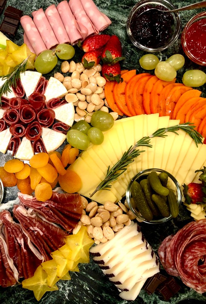 A selection of carefully arranged sliced cheeses, cured meats, dried and fresh fruit, nuts, jams, cornichon, chocolates, and rosemary sprigs.