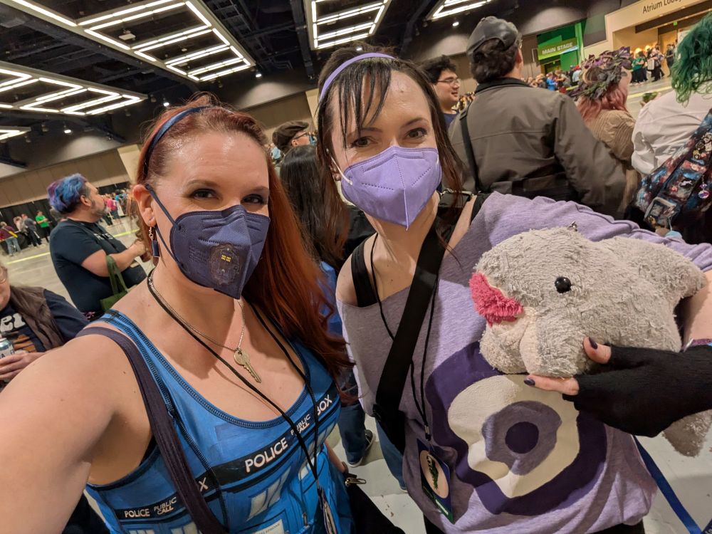 Lorelei and Rose from the waist up. Lorelei is wearing a blue headband over long red hair over one shoulder, blue mask with a Tardis sticker on one side, key necklace, Tardis tank top dress, and lanyard badge, next to Rose, wearing a light purple headband over black hair, a purple mask, a light purple tee shirt with a purple and white bullseye, and black gauntlets, holding Jeff, a plush grey shark with feet.