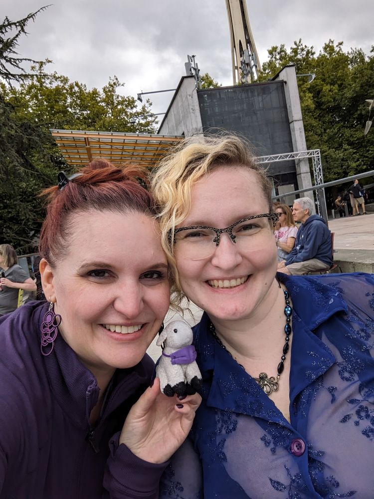Lorelei with her red hair in a messy bun, wearing big purple earrings and a purple shirt, holding Sheepy the tiny stuffed sheep with his purple mask around his neck, affectionately head to head with Rose, with bleached blonde bangs, wearing glasses, an octopus necklace, and a sheer purply-blue collared shirt, outdoors at Seattle Center.