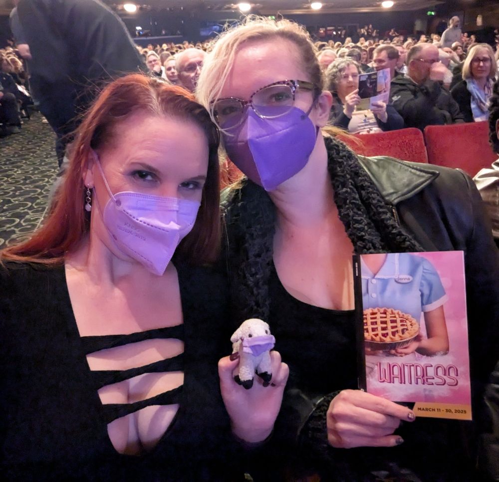 Lorelei with red hair wearing a purple mask and a cleavage-revealing black sweater, Rose with blonde bangs wearing glasses, a purple mask, a black leather jacket, and a black shirt, Sheepy the tiny stuffed sheep wearing a purple mask, and the playbill for the musical Waitress, with the theater crowd in the background.