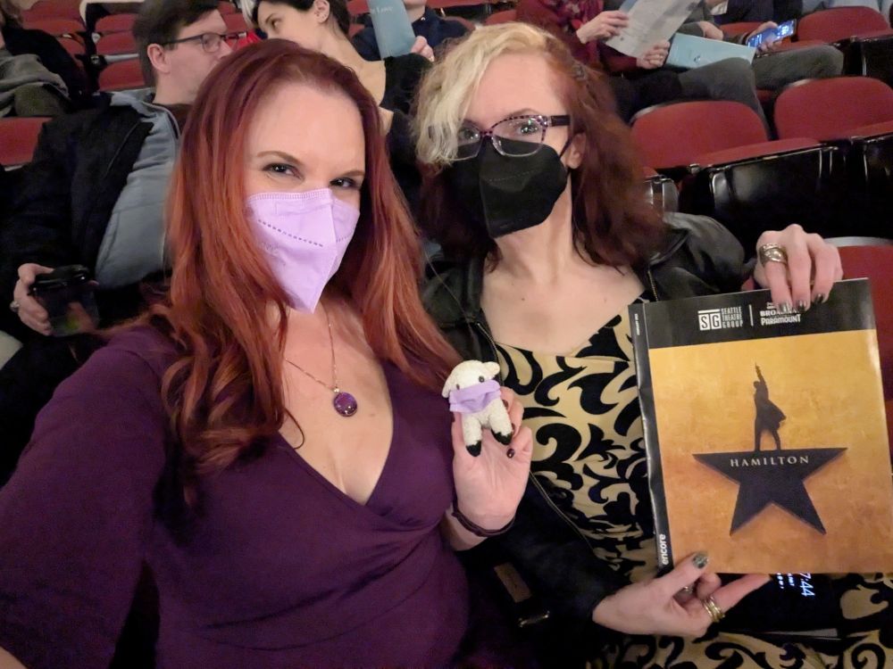 Lorelei with long red hair cascading down her shoulders, wearing a low-cut purple dress, a purple mask, and a purple pendant, holding Sheepy the tiny stuffed sheep in his own purple mask, with Rose with red and blonde hair, a black mask, and a black and cream floral dress, holding the playbill for Hamilton. There are red theater seats and other patrons in the background.