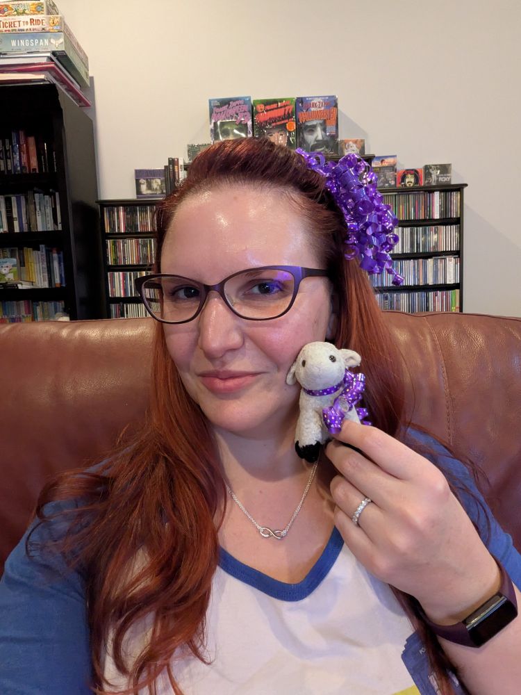 Lorelei from the bust up wearing purple glasses, a blue and white TARDIS T-shirt, silver infinity necklace, a purple watch, and purple curled holiday ribbons as a hair accessory, holding Sheepy the tiny stuffed sheep, wearing his own purple curled holiday ribbon as a jaunty scarf. Mr Rivers's enormous CD collection is in the background.