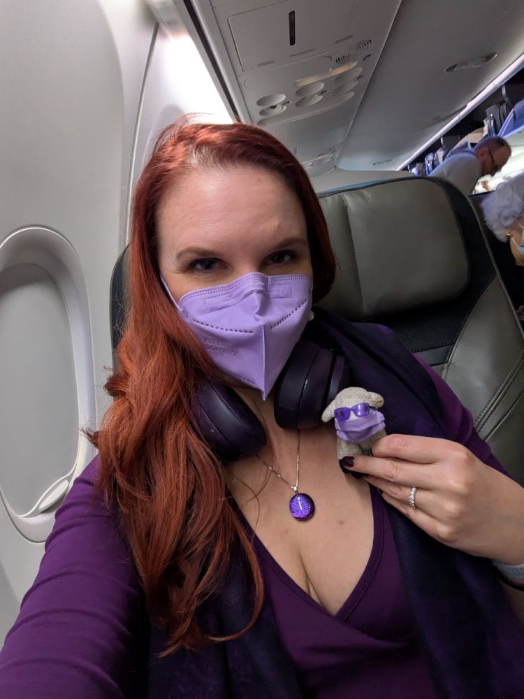 Lorelei with red hair cascading down one shoulder, wearing a purple mask, purple pendant, and low-cut purple dress, holding Sheepy the tiny stuffed sheep, who is wearing his own purple mask and purple sunglasses, in an airplane seat