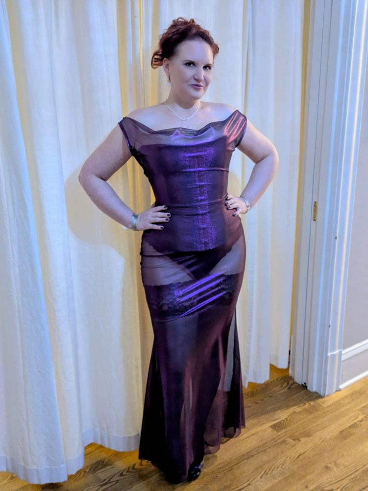 A full length photo of Lorelei, smiling mischievously, posing with hands on hips, with an elegantly curled updo hairstyle, wearing a tight laced overbust corset, black panties, black stockings, and very high heels under a sheer shimmery off-shoulder dress, and accessorized with silver infinity necklace, silver bracelets, and purple polished fingernails.