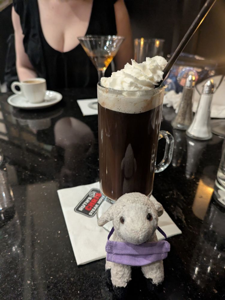 Sheepy the tiny stuffed sheep with his purple mask around his neck, with a whipped cream-topped coffee in a glass mug behind him on a Ruth's Chris Steak House napkin, and a martini, a tiny espresso, and Rose's decolletage in the background