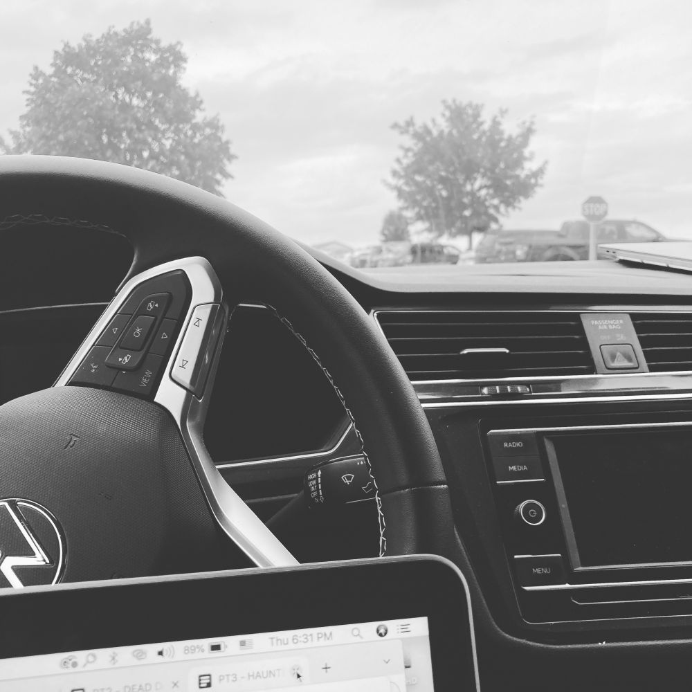 The steering wheel and dashboard of a car with a bit of a laptop computer. Two trees are seen in the distance.