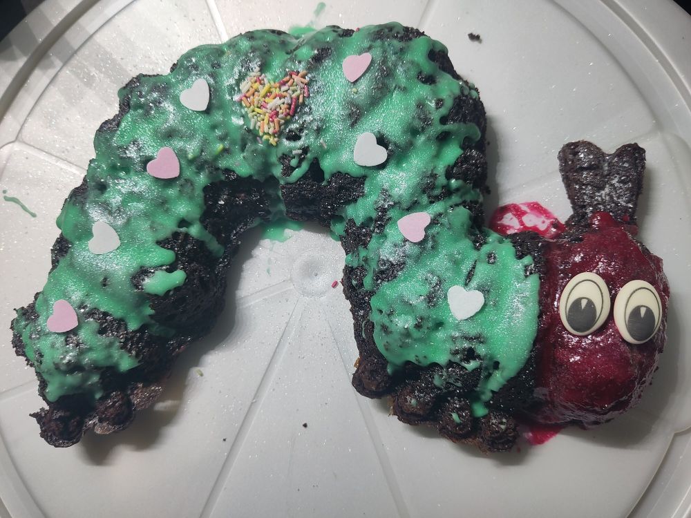 Eine Raupe Nimmersatt aus Schokokuchen, mit grünem und rotem Zuckerguss, Zuckeraugen und essbarem Glitzer. In der Mitte des Körpers ist ein Herz aus bunten Streuseln und auf dem Rest der Raupe sind rosa und weiße Esspapierherzen verteilt.