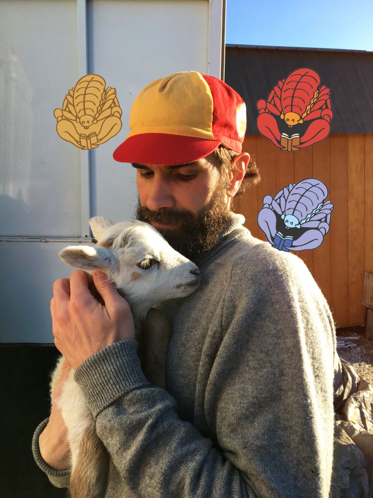 Pin artist Daniel Mrgan holding a baby goat outside surrounded by pseudoscorpion pin designs. 