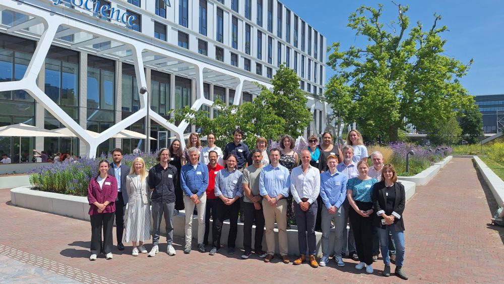A group of 25 people standing together in front of a building to the left and tree to the right