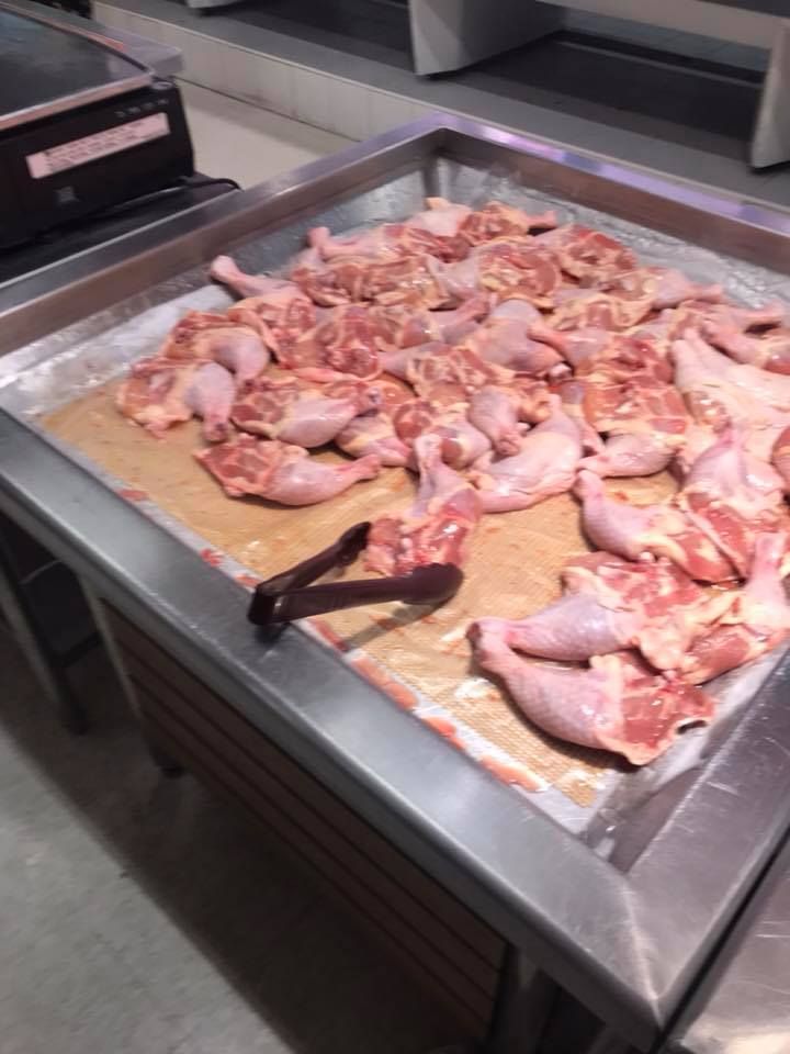 Auf dem Bild ist ein offenes Fleischregal in einem Supermarkt zu sehen. Darauf liegen viele rohe Hähnchenschenkel nebeneinander auf einer flachen Metallfläche, die mit einer Unterlage ausgelegt ist. Vorne liegt eine Zange, mit der Kunden die Hähnchenteile aufnehmen können. Im Hintergrund ist eine Waage zu erkennen, vermutlich zum Abwiegen des Fleisches. Das gesamte Setting wirkt wie eine Frischetheke im Selbstbedienungsbereich.