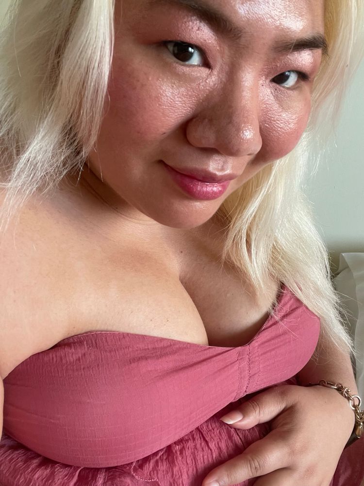 A blonde Asian non-binary woman is holding their pregnant belly with their left hand. They are wearing a rose dress and rose makeup.
