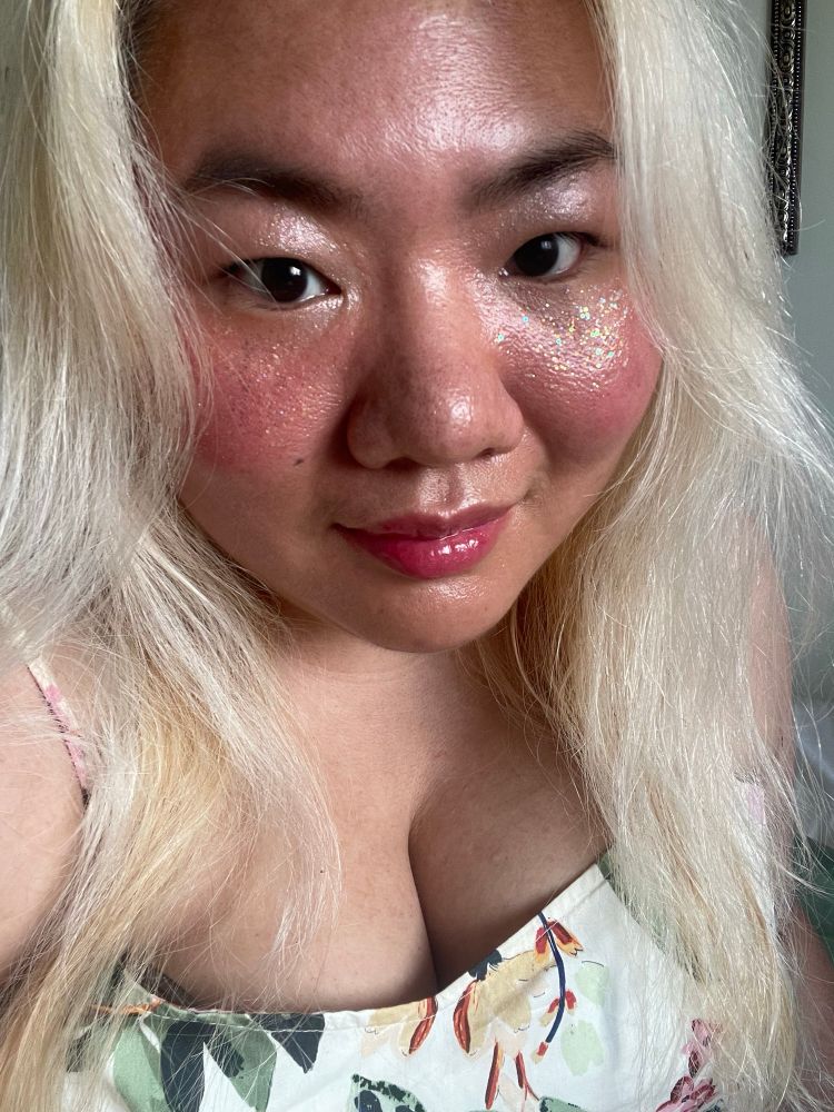 A blonde non-binary Asian woman smiles at the camera. They are wearing light makeup: lip gloss and body glitter.