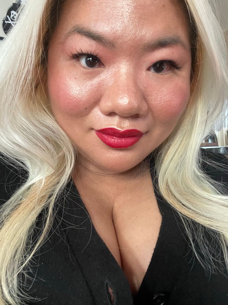 A blonde Asian non-binary woman smiles at the camera. They are wearing a full face of light makeup with a bright red lip. They are also wearing a black cardigan that shows off their deep cleavage.