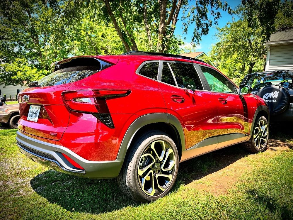 The right rear three-quarter view of a 2025 Chevrolet Trax 2RS in Crimson Red parked in a driveway under the midday sun behind a black 1997 Toyota RAV4 