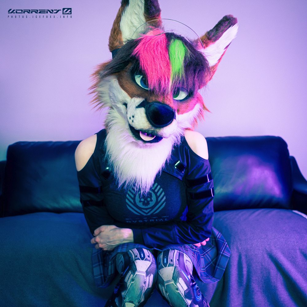 A photo of me wearing my fox fursuit head, a shrug by Crisiswear, a tank top by Dustrial, a pleated skirt, and leggings with a robot legs print on them. I'm sitting on a black couch leaning slightly towards the camera.