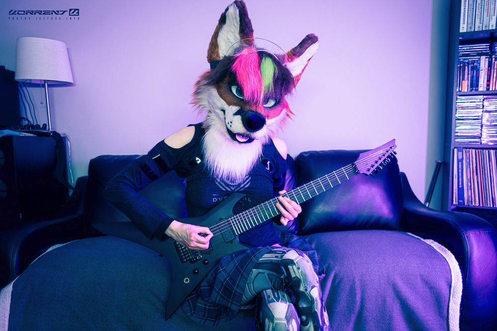 A photo of me wearing my fox fursuit head, a shrug by Crisiswear, a tank top by Dustrial, a pleated skirt, and leggings with a robot legs print on them. I'm sitting on a black couch holding my Ibanez Xiphos 7-string guitar and looking into the camera.
