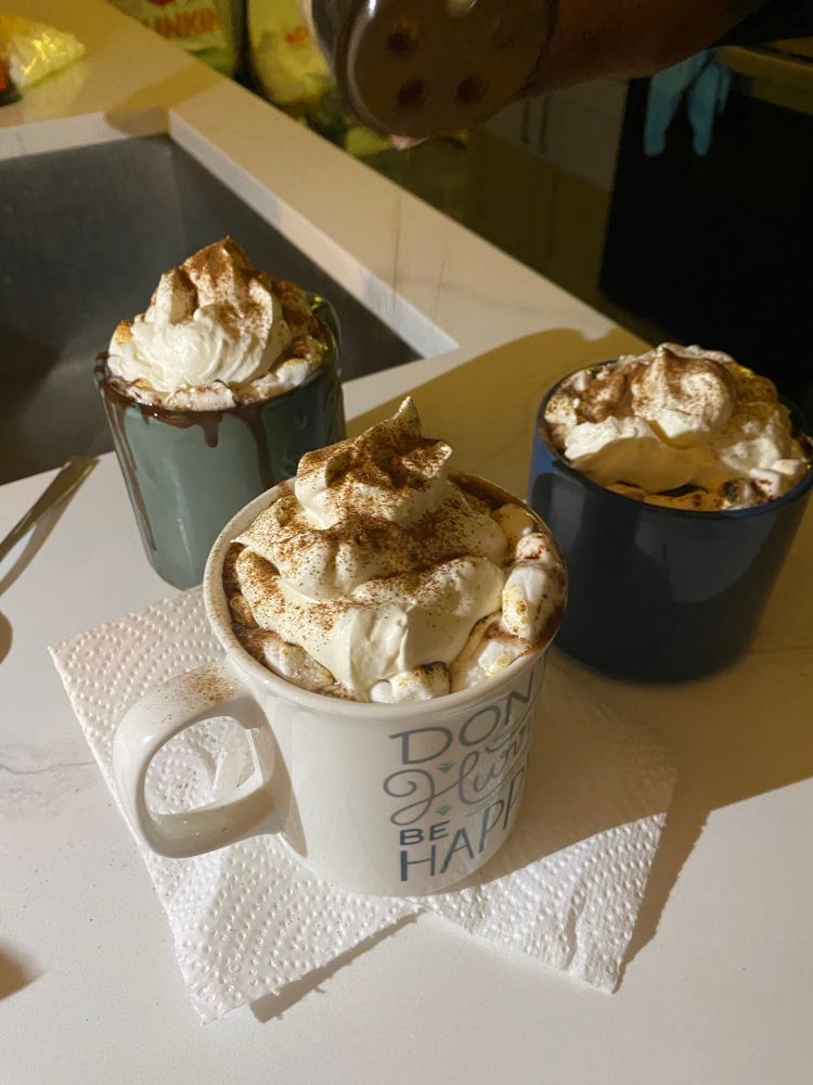 Three mugs of hot chocolate on a counter. There are marshmallows and whipped cream
