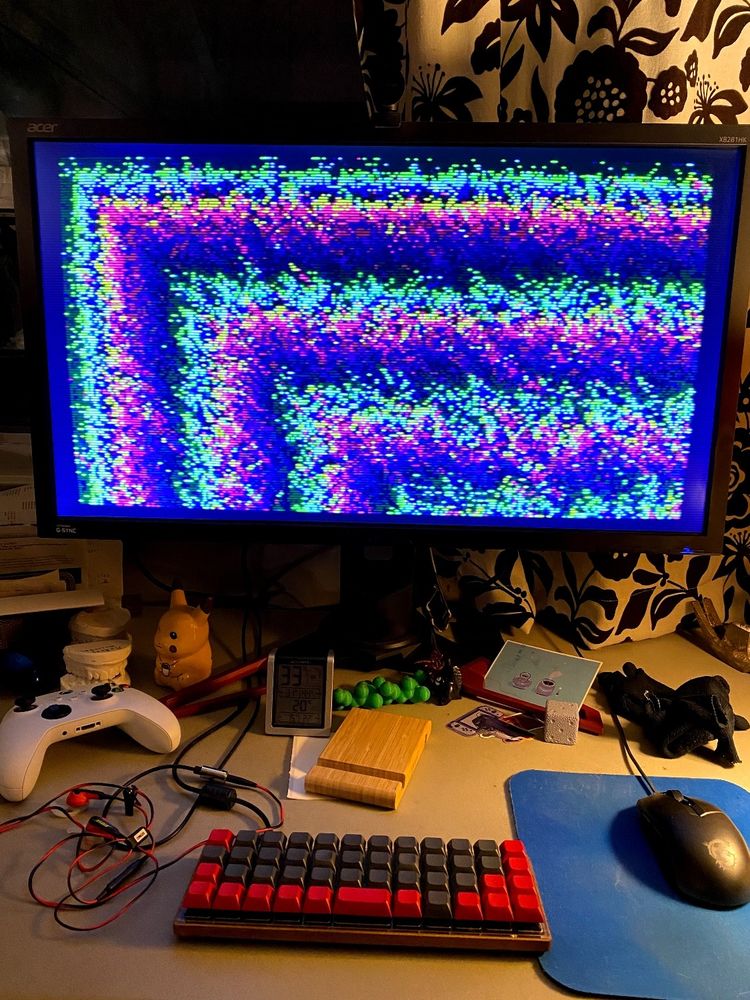 A desk, monitor shows some abstract generative pixelart. There's a tiny Planck keyboard.