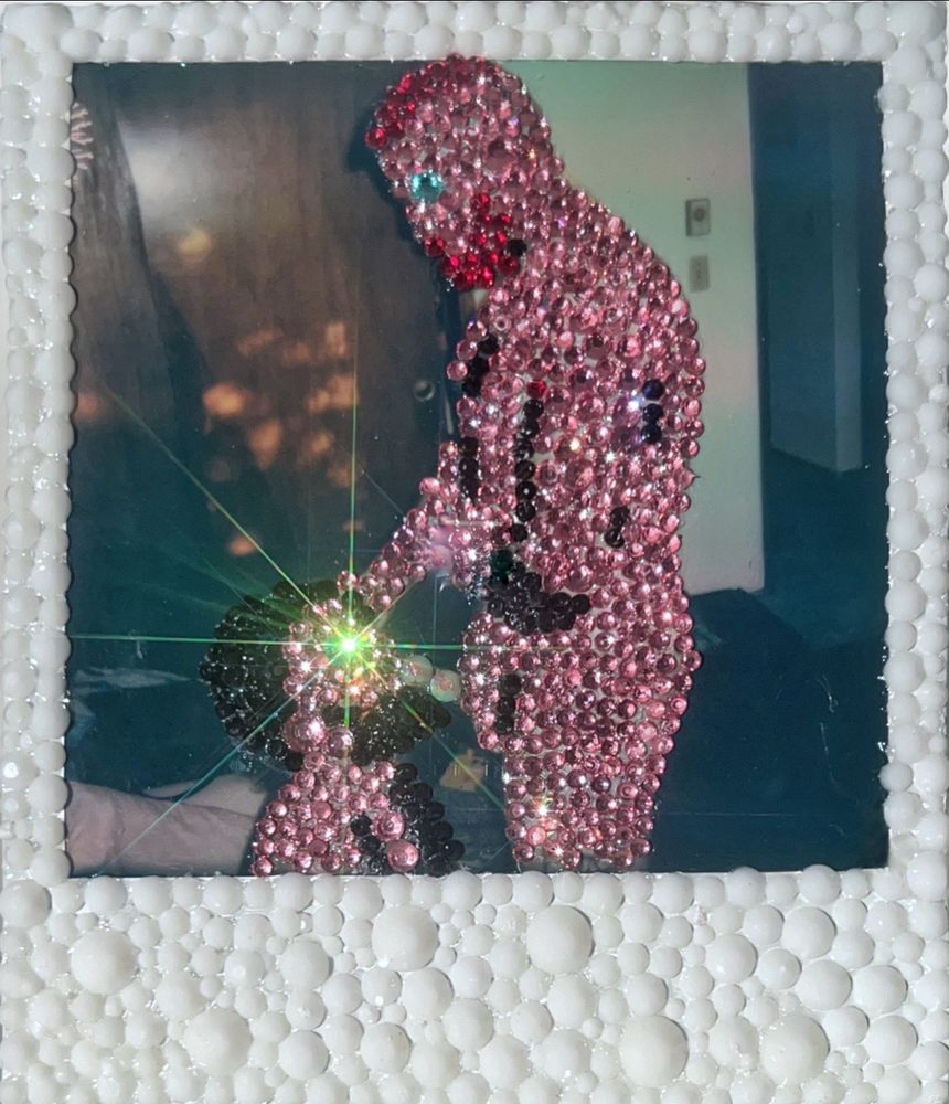 A Polaroid of a man performing oral sex on another has been meticulously covered in rhinestones. The people are fully covered in stones creating a humanoid effect and have both been anonymized in the process. The backdrop has been left un-stoned, making the figures pop out in an eye catching way. A star shaped lense flair emanates from the eye of the man on his knees. 
White rhinestones adorn the entire white border of the Polaroid. 