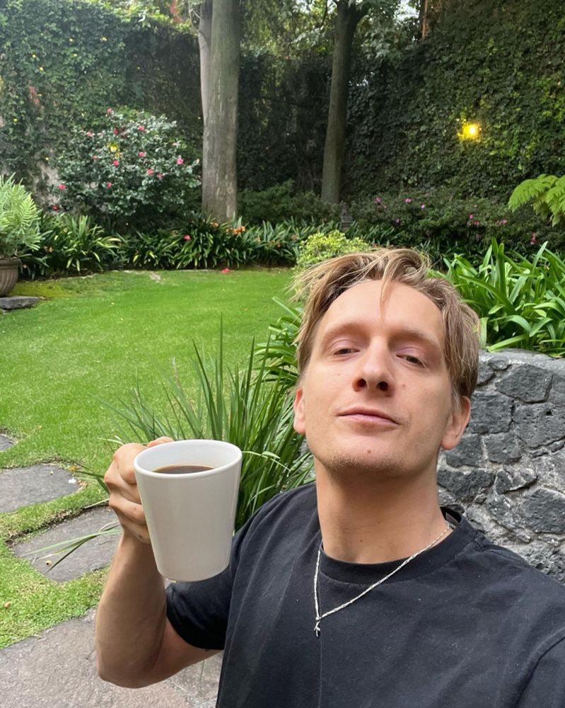 Auf dem Bild sieht man Felix Kummer, der entspannt in einem Garten steht und eine Tasse schwarzen Kaffee in der rechten Hand hält. Die Person trägt ein schwarzes T-Shirt und eine silberne Kette mit einem kleinen Anhänger. Sie hat blonddunkelblonde, leicht zerzauste Haare und schaut selbstbewusst in die Kamera mit einem leichten Lächeln.

Im Hintergrund ist ein gepflegter Garten mit einem satten grünen Rasen, verschiedenen Pflanzen, Blumen und hohen Bäumen zu sehen. Eine Steinmauer und ein kleiner Steinweg ergänzen die idyllische, ruhige Atmosphäre. Es wirkt wie ein entspannter Morgenmoment mit Kaffee im Grünen. 🌿☕