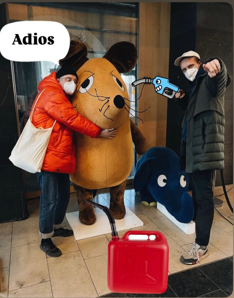 Das Bild zeigt zwei Personen, Steffen Israel und Felix Kummer von der Band Kraftklub, mit Masken, die neben einer großen Stofffigur der bekannten deutschen Kinderfernsehfigur „Die Maus“ stehen. Die linke Person trägt eine rote Daunenjacke und umarmt die Maus, während eine Sprechblase mit dem Text „Adios“ erscheint. Die rechte Person trägt eine dunkle Jacke und zeigt auf die Kamera. Im Hintergrund ist auch der Elefant aus der „Sendung mit der Maus“ zu sehen. Humorvoll überarbeitet wurde das Bild mit einem gezeichneten Benzinschlauch, der in die Maus gesteckt ist, sowie einem roten Benzinkanister am Boden. Der gezeichnete Schlauch kommt von der rechten Person, was eine ironische oder sarkastische Szene andeutet.
