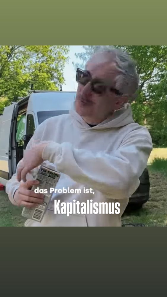 Steffen Israel steht draußen vor einem weißen Van auf einer Wiese, trägt eine helle Kapuzenjacke und hält einen Getränkekarton in der Hand. Er trägt eine dunkle Sonnenbrille und gestikuliert mit einem leicht ironischen Ausdruck. Im Bild ist der Text eingeblendet: „das Problem ist, Kapitalismus“.